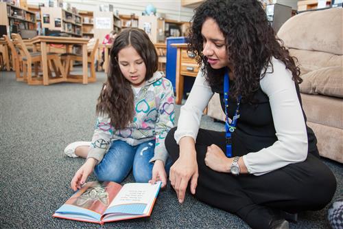 School administrator and student read a book together in the library.  
